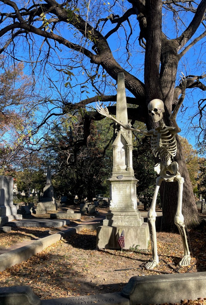 A larger than life size skeleton posed with arms raised in front of an obelisk grave in the Woodlands cemetery in Philadelphia